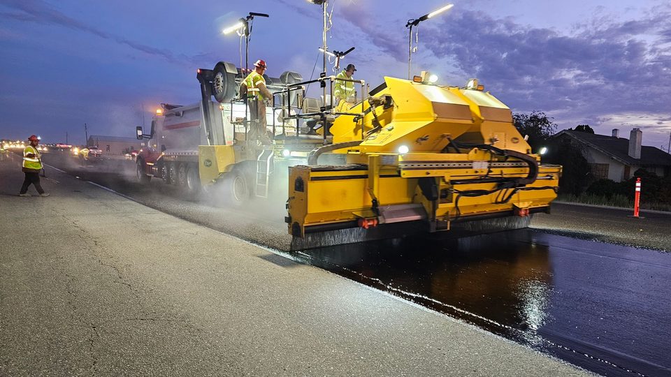 Paving crew laying a fresh asphalt mat behind a paver under work lights