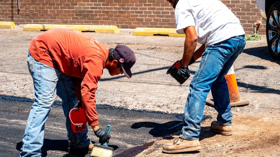 Two workers hand-finishing an asphalt patch along a concrete curb line
